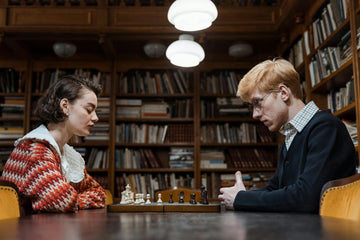 A woman sits across from a man playing chess with a shelf of books in the background.